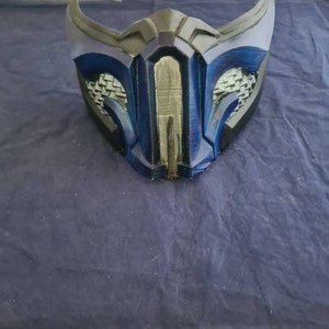 May include: A blue and black plastic mask with a geometric design. The mask has a raised, textured surface and a small opening for the mouth.