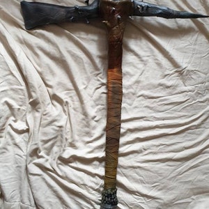 May include: A dark brown and black spiked weapon with a long, wrapped handle. The handle is wrapped in brown, yellow, and orange fabric. The weapon has a pointed tip and a decorative metal top.
