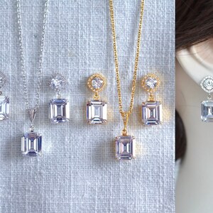 May include: A set of emerald cut cubic zirconia earrings and a pendant necklace. The earrings and pendant feature a smaller round cubic zirconia halo. The necklace and earrings are available in gold and silver.
