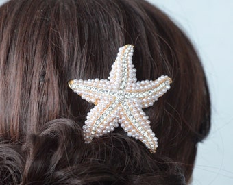 May include: A gold starfish hair clip with white and pink pearl accents. The clip is worn in a woman's dark brown hair.