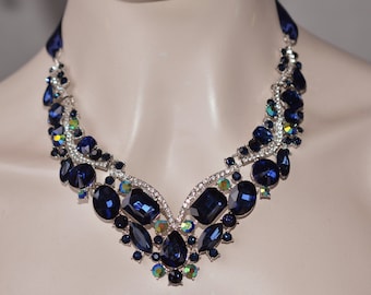 May include: A navy blue statement necklace with large, faceted rhinestones. The necklace is made of silver-toned metal and features a V-shaped design.