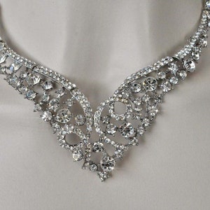 May include: A silver necklace with a delicate, intricate design featuring a cluster of clear rhinestones. The necklace has a V-shaped neckline and is perfect for a formal occasion.