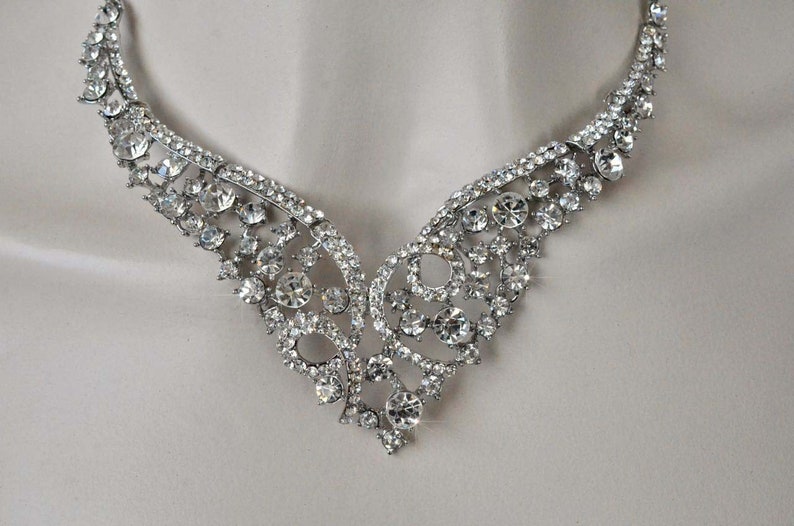 May include: A silver necklace with a delicate, intricate design featuring a cluster of clear rhinestones. The necklace has a V-shaped neckline and is perfect for a formal occasion.