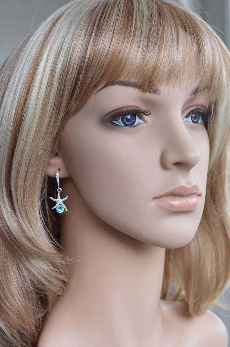 Puede incluir: Un maniqu&iacute; con cabello rubio que lleva un par de aretes colgantes de plata con una piedra preciosa azul y un dije de estrella de mar.