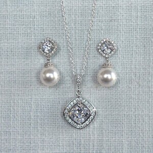 May include: A silver necklace and earrings set with a large, clear, square-shaped cubic zirconia pendant and matching earrings. The pendant and earrings are surrounded by smaller cubic zirconia stones. The necklace has a delicate silver chain and the earrings have a pearl drop.
