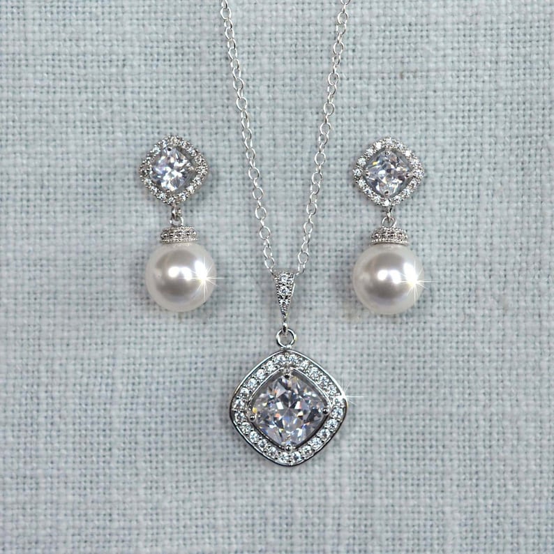 May include: A silver necklace and earrings set with a large, clear, square-shaped cubic zirconia pendant and matching earrings. The pendant and earrings are surrounded by smaller cubic zirconia stones. The necklace has a delicate silver chain and the earrings have a pearl drop.