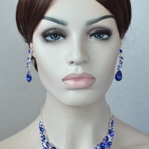 May include: A silver necklace and earrings set with teardrop-shaped blue rhinestones. The necklace features a cascading design with multiple layers of rhinestones.