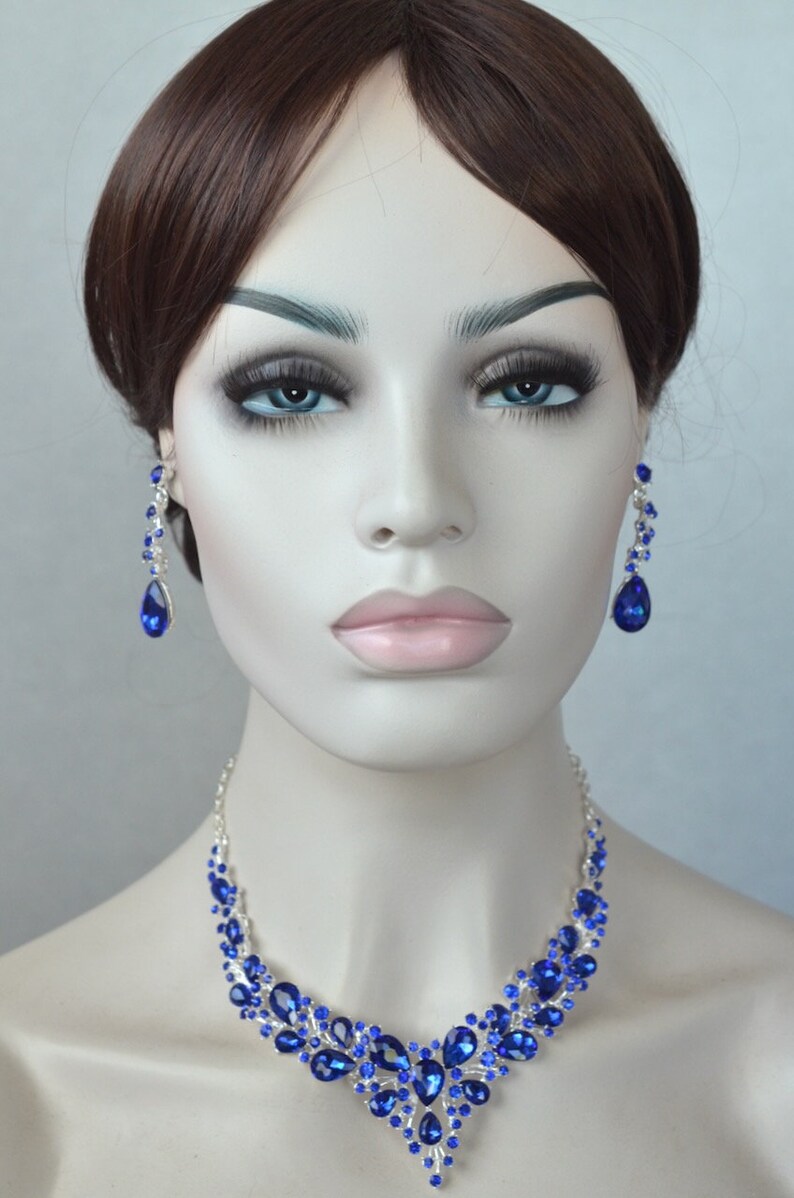 May include: A silver necklace and earrings set with teardrop-shaped blue rhinestones. The necklace features a cascading design with multiple layers of rhinestones.