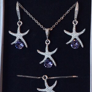 Puede incluir: Un conjunto de collar y pendientes de plata con un dise&ntilde;o de estrella de mar y un cristal morado en el centro. El collar y los pendientes se muestran en una caja blanca.