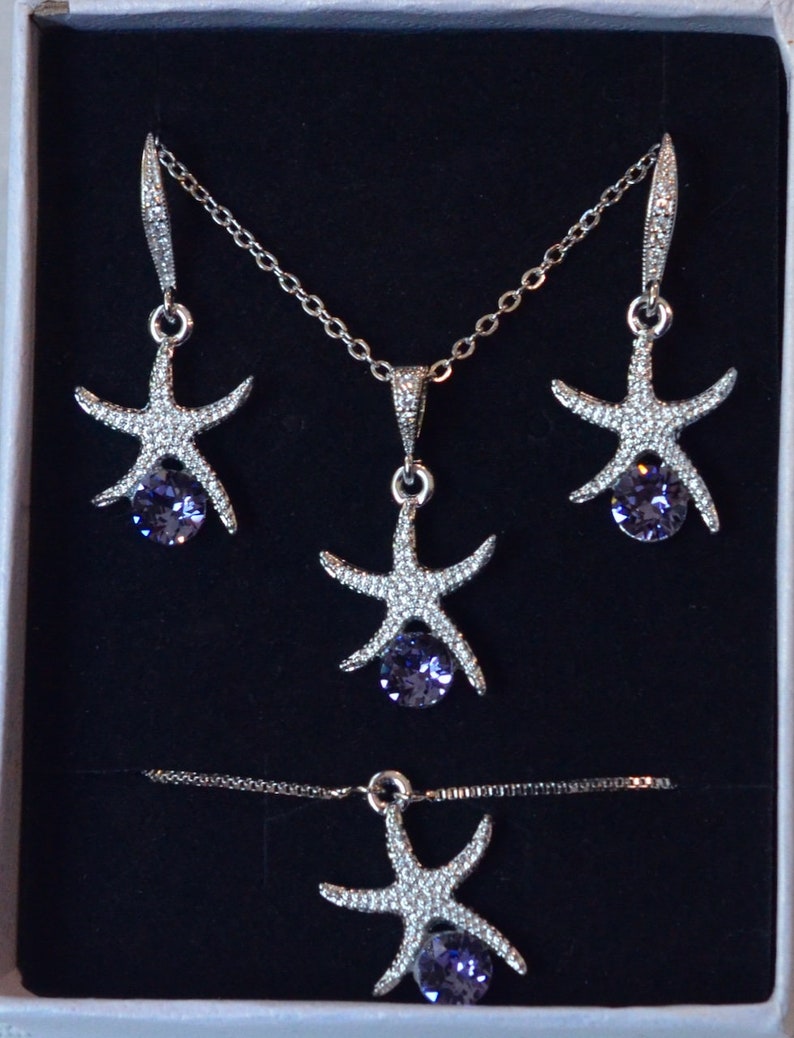 Puede incluir: Un conjunto de collar y pendientes de plata con un dise&ntilde;o de estrella de mar y un cristal morado en el centro. El collar y los pendientes se muestran en una caja blanca.