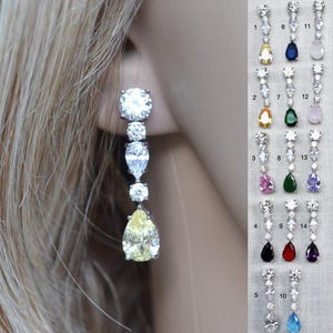 May include: A long drop earring featuring a silver-tone setting with three round clear stones and a large pear-shaped yellow stone at the base. A vertical display shows fourteen numbered variations of this earring with different colored stones.