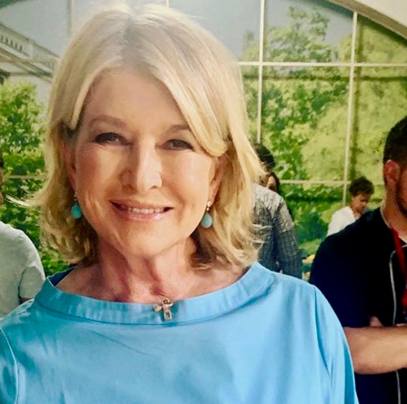 May include: A woman with blonde hair is wearing a light blue blouse with a small silver pendant necklace. She is wearing turquoise teardrop earrings.