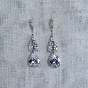 May include: A pair of silver dangle earrings with teardrop-shaped crystals and smaller crystals clustered around the top of the teardrop.