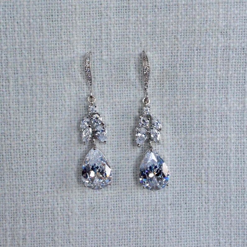 May include: A pair of silver dangle earrings with teardrop-shaped crystals and smaller crystals clustered around the top of the teardrop.