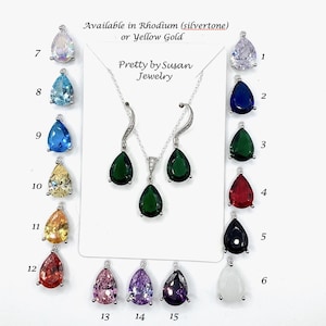 May include: A jewelry set featuring a necklace and earrings with dark green pear-shaped stones on a display card labeled Pretty by Susan Jewelry. Surrounding the card are fifteen numbered pear-shaped pendants in various colors including clear, blue, green, red, black, white, yellow, orange, pink, and purple.