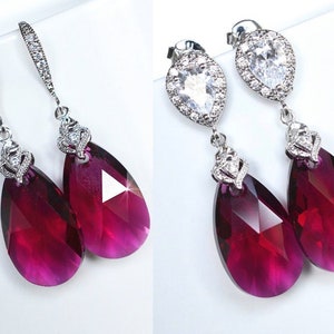 May include: A pair of dangle earrings featuring a teardrop-shaped, faceted, deep purple crystal. The crystal is surrounded by a silver setting with a smaller clear crystal halo. The earrings have a silver hook fastening.