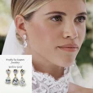 May include: Elegant yellow gold earrings with a pear-shaped cubic zirconia stone. The earrings are displayed on a white card with the text "Pretty by Susan Jewelry" and "Yellow Gold". The earrings are shown on a person.