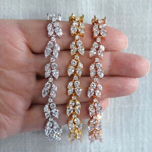 May include: Three delicate bracelets with a leaf design, each with a different metal finish: silver, gold, and rose gold. The bracelets are adorned with sparkling cubic zirconia stones.