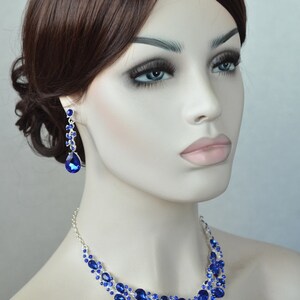 May include: A silver necklace and earrings set with blue crystal stones. The necklace features a teardrop design with a cascading pattern of smaller stones. The earrings are long and dangle with a teardrop stone at the bottom.