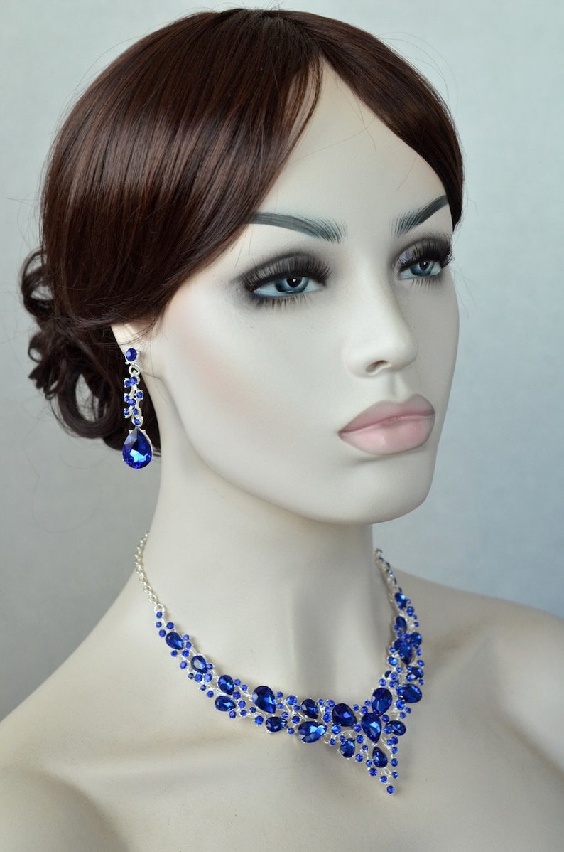 May include: A silver necklace and earrings set with blue crystal stones. The necklace features a teardrop design with a cascading pattern of smaller stones. The earrings are long and dangle with a teardrop stone at the bottom.