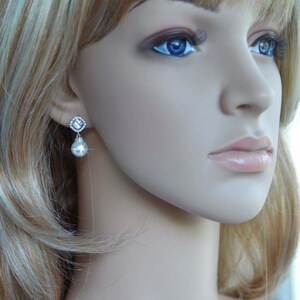 May include: A pair of silver earrings with a clear crystal and a pearl drop. The earrings are on a mannequin with blonde hair.