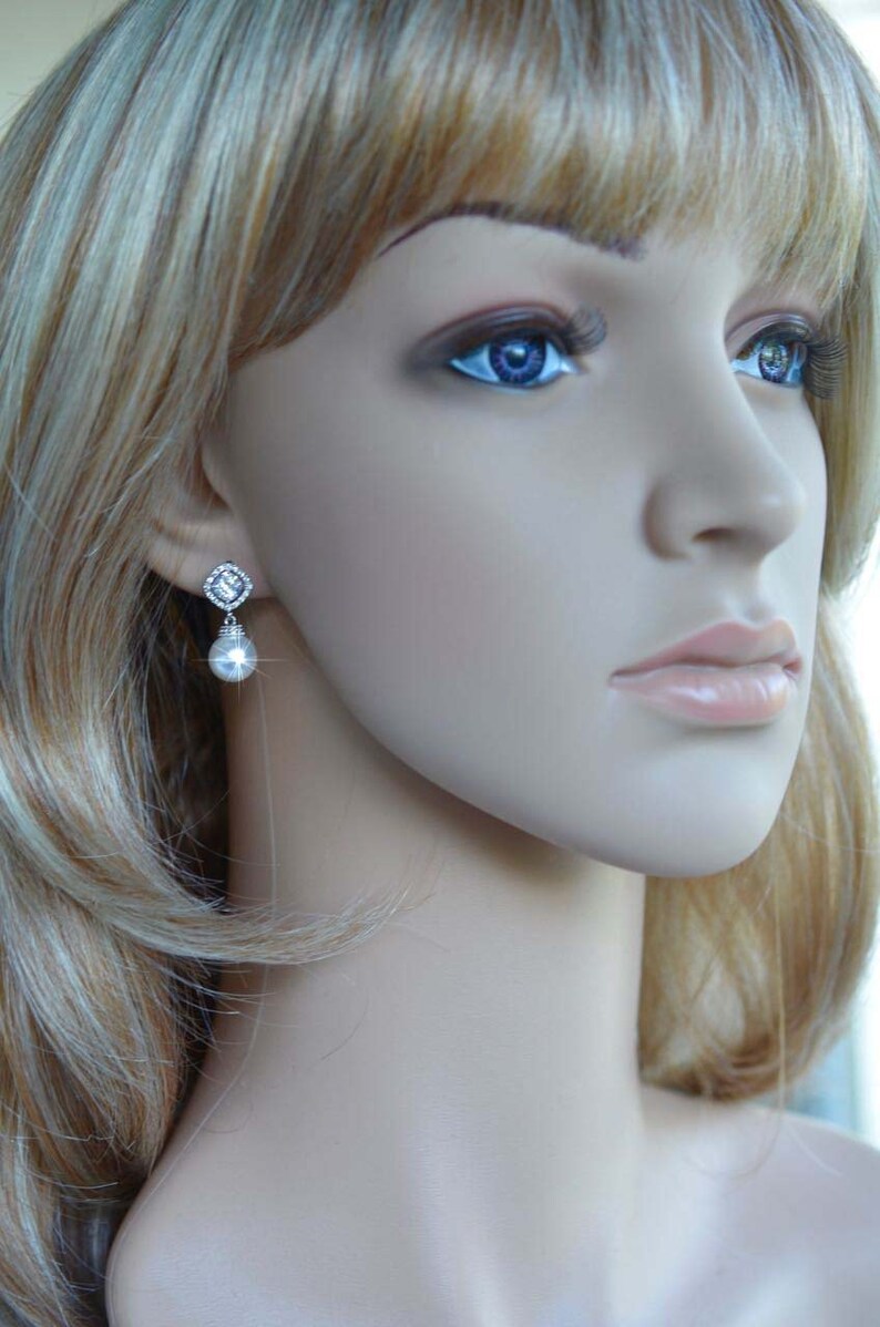 May include: A pair of silver earrings with a clear crystal and a pearl drop. The earrings are on a mannequin with blonde hair.