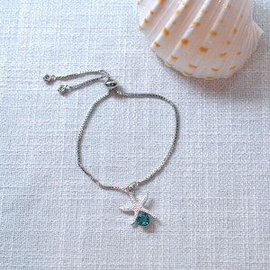 Puede incluir: Un brazalete ajustable de plata con un dije de estrella de mar con un cristal azul turquesa.