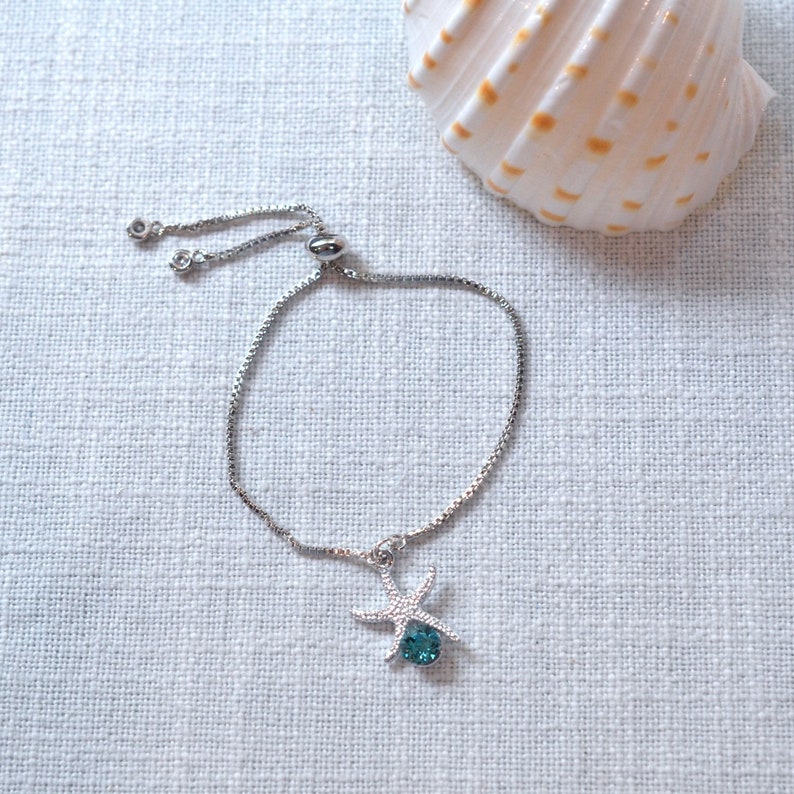 Puede incluir: Un brazalete ajustable de plata con un dije de estrella de mar con un cristal azul turquesa.