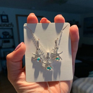 Puede incluir: Un conjunto de collar y pendientes de plata con un dise&ntilde;o de estrella de mar y cristales iridiscentes. Los colgantes de estrella de mar est&aacute;n detallados con peque&ntilde;as piedras brillantes.