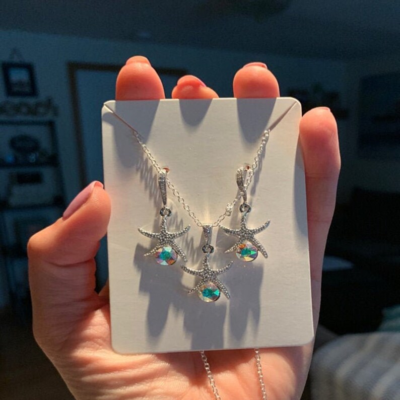 Puede incluir: Un conjunto de collar y pendientes de plata con un dise&ntilde;o de estrella de mar y cristales iridiscentes. Los colgantes de estrella de mar est&aacute;n detallados con peque&ntilde;as piedras brillantes.