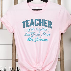 Op de afbeelding: Een zachtroze t-shirt met de tekst "TEACHER of the brightest 2nd Grade Stars Mrs Johnson" in blauw en teal. Het shirt hangt aan een zwarte hanger tegen een witte achtergrond.