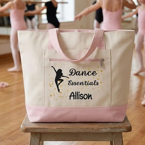 Puede incluir: Bolso tote de lona color crema con detalles en rosa, bolsillo frontal y asas. El bolso muestra una silueta negra de bailarina, las palabras "Dance Essentials" y el nombre "Allison" en dorado. El bolso está sobre un taburete de madera.