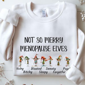 Op de afbeelding: Witte sweatshirt met de zwarte tekst "NOT SO MERRY MENOPAUSE ELVES". Daaronder illustraties van elfen met de woorden: Itchy, Bitchy, Bloated, Sleepy, Sweaty, Forgetful en Psyc'.