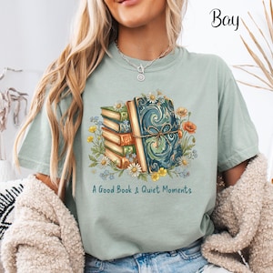 May include: A light green t-shirt featuring a stack of books and floral illustrations. The design includes a large, ornate book tied with a string, surrounded by daisies, poppies, and other flowers. The text "A Good Book & Quiet Moments" is printed below.
