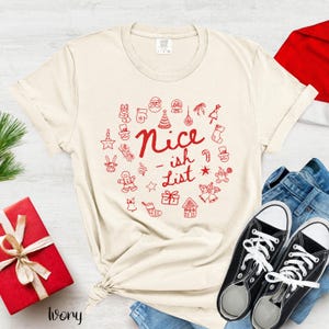 May include: Cream-coloured t-shirt featuring a red "Nice-ish List" design surrounded by festive doodles. The shirt is paired with black trainers and blue jeans, and a red gift box with a bow.