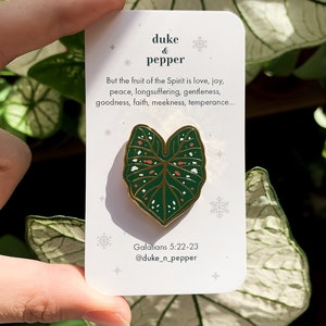 May include: A green and gold enamel pin in the shape of a leaf, with white and orange details, is displayed on a white card. The card has text that reads "duke & pepper" and a Bible verse. The pin is held by a hand.