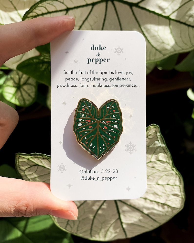 May include: A green and gold enamel pin in the shape of a leaf, with white and orange details, is displayed on a white card. The card has text that reads "duke & pepper" and a Bible verse. The pin is held by a hand.