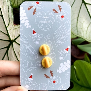 May include: Two yellow pin backs on a light gray card with a repeating pattern of dog and leaf illustrations. The dogs are wearing Santa hats and reindeer antlers. The text "duke & pepper" is also on the card.