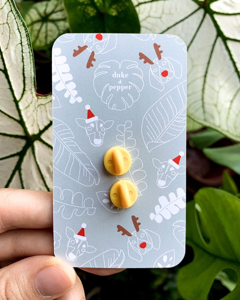 May include: Two yellow pin backs on a light gray card with a repeating pattern of dog and leaf illustrations. The dogs are wearing Santa hats and reindeer antlers. The text "duke & pepper" is also on the card.