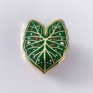 May include: A gold-edged enamel pin in the shape of a green leaf. The leaf has white and red accents, with a detailed vein pattern. The pin is a botanical accessory.