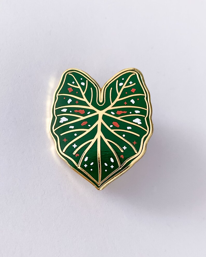 May include: A gold-edged enamel pin in the shape of a green leaf. The leaf has white and red accents, with a detailed vein pattern. The pin is a botanical accessory.