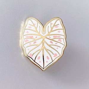 May include: A gold-colored metal pin in the shape of a leaf. The leaf is white with pink spots and gold veins. The pin has a gold border and star-shaped accents.