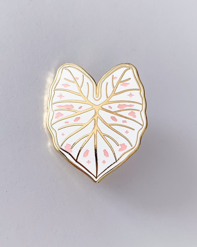 May include: A gold-colored metal pin in the shape of a leaf. The leaf is white with pink spots and gold veins. The pin has a gold border and star-shaped accents.
