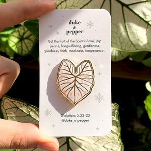 May include: A gold-colored enamel pin in the shape of a leaf with pink accents, held against a white card. The card features text that reads "duke & pepper" and a Bible verse. The pin is a decorative accessory.