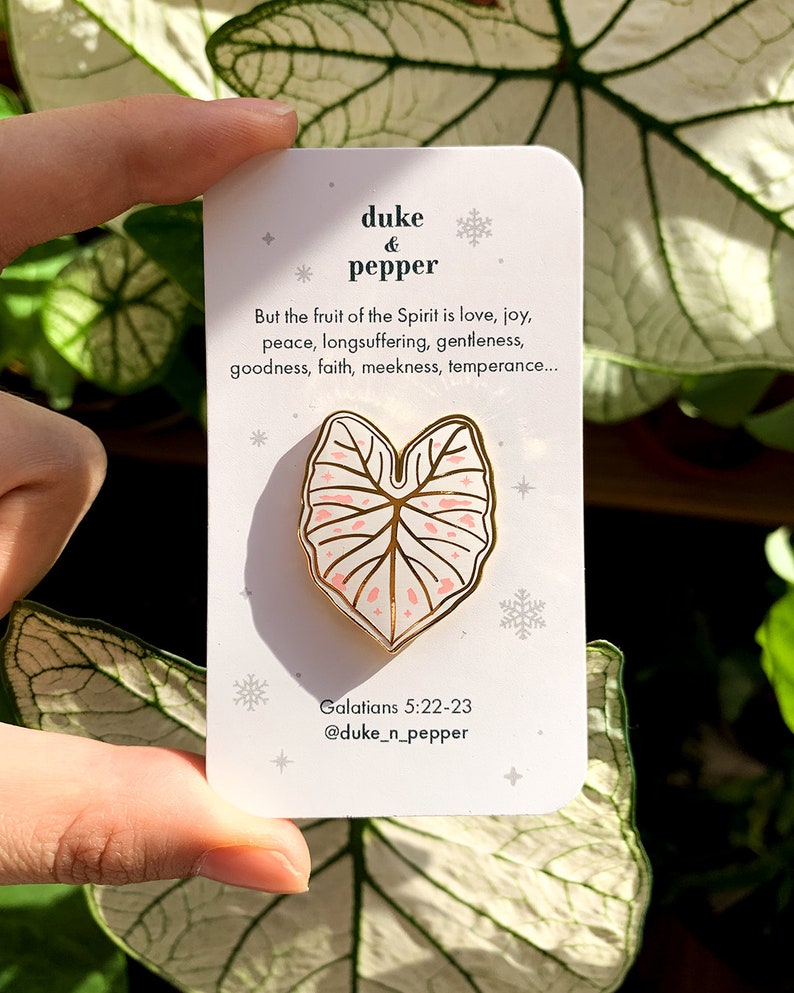 May include: A gold-colored enamel pin in the shape of a leaf with pink accents, held against a white card. The card features text that reads "duke & pepper" and a Bible verse. The pin is a decorative accessory.
