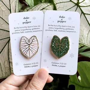 May include: Two enamel pins featuring leaf designs, one pink and white, the other green and white, with gold-colored borders. Each pin is on a white card with text that reads "duke & pepper" and a Bible verse. The pins are held by a hand.