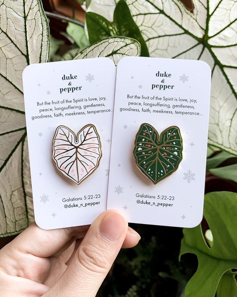 May include: Two enamel pins featuring leaf designs, one pink and white, the other green and white, with gold-colored borders. Each pin is on a white card with text that reads "duke & pepper" and a Bible verse. The pins are held by a hand.