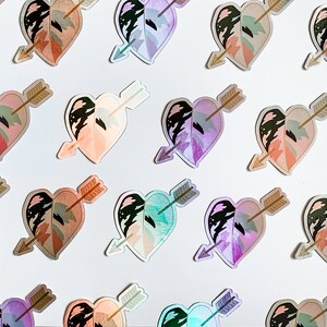 May include: A collection of heart-shaped stickers featuring a leaf design pierced by an arrow. The stickers have a holographic effect, with colors ranging from pink and peach to green and purple. The arrow is a light brown color.