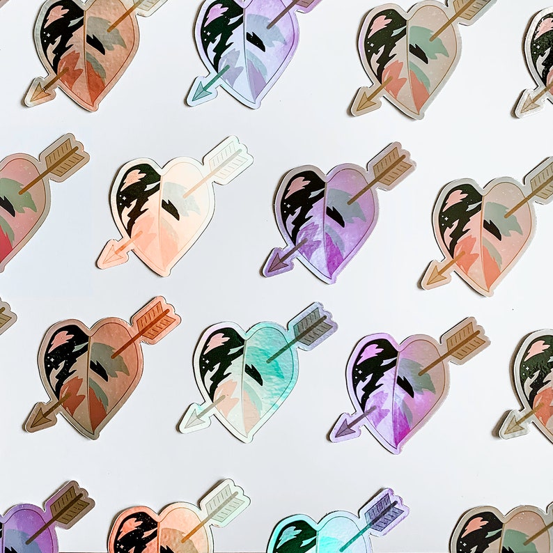 May include: A collection of heart-shaped stickers featuring a leaf design pierced by an arrow. The stickers have a holographic effect, with colors ranging from pink and peach to green and purple. The arrow is a light brown color.