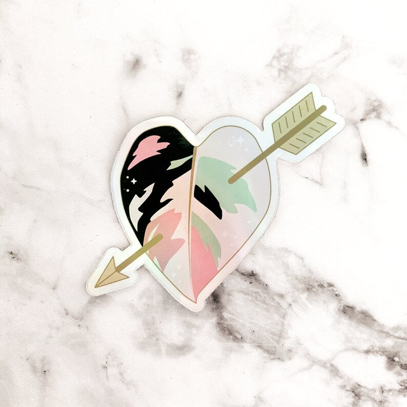 May include: A heart-shaped sticker with a leaf design, bisected by a gold arrow. The leaf features pink, black, and green sections. The sticker has a holographic, iridescent finish, creating a shimmering effect.